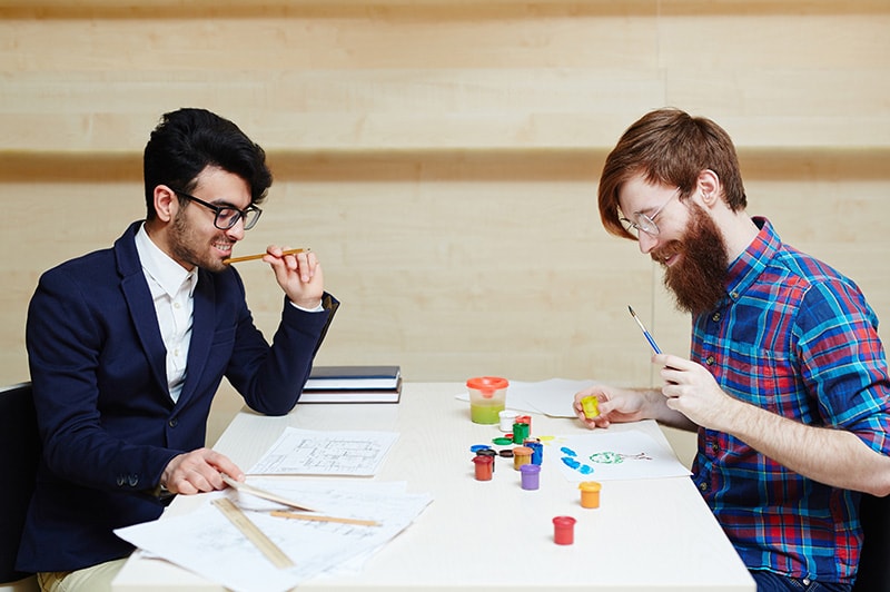 Side view of two bearded men in eyeglasses: one of them wearing suit and white shirt making changes to blueprint while the other drawing picture with brush and gouache