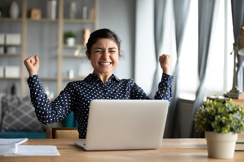 Overjoyed young Indian woman sit at desk in living room feel euphoric win lottery online on laptop, excited millennial ethnic girl triumph receive read pleasant approval email on computer