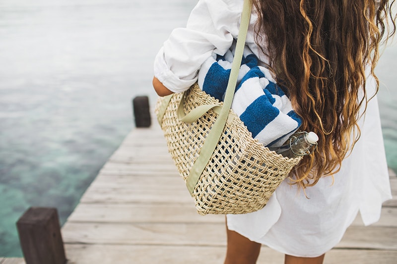 Woman with handmade wicker bag, two beach towels and glass bottle for water going to the beach. View from behind close up. Eco friendly and zero waste concept.